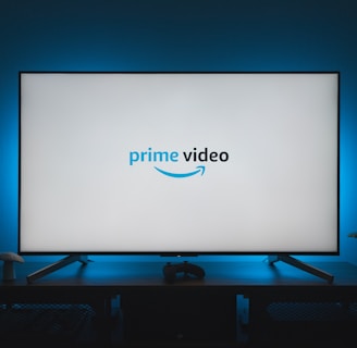 A television screen displays the Prime Video logo, situated on an entertainment unit in a dimly lit room. The room is illuminated with a subtle blue backlight, creating a calming atmosphere. Two small decorative items, resembling mushrooms, are placed on either side of the unit.